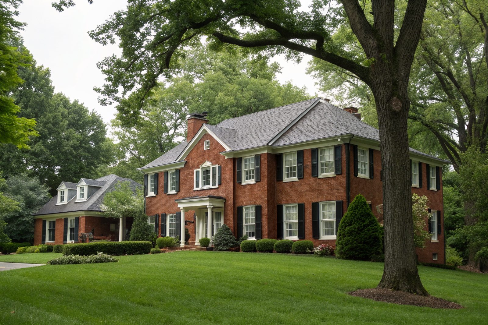 A two-story red brick colonial home with a charcoal shingle roof, black shutters and mature oaks